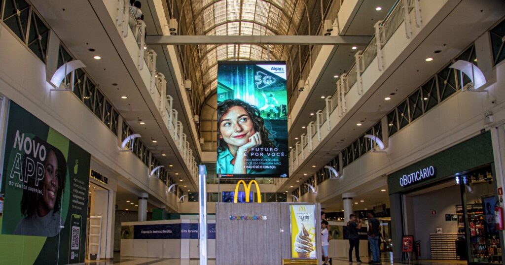 Imagem do corredor central de um shopping center com destaque de um painel de LED suspenso exibindo publicidade. 