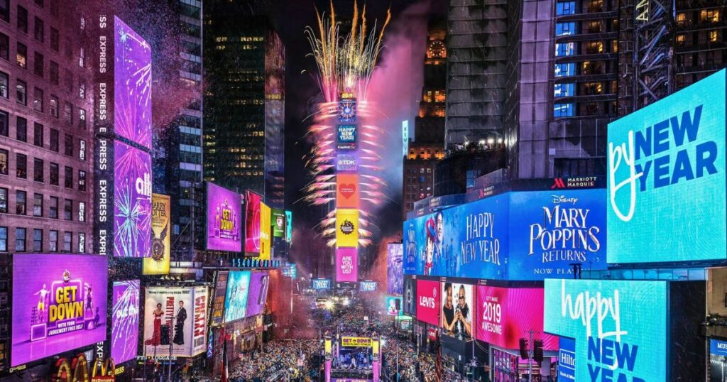Imagem mostra o momento exato da virada de ano na contagem regressiva da Times Square com o relógio e a famosa bola de LED ao fundo, a rua coberta por pessoas e os prédios todos iluminados com anúncios de teatro e publicidade em painéis de LED.
