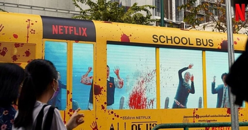 Imagem mostra uma das ações publicitárias outdoor da Netflix para promover o k-drama All of Us are Dead. Na imagem pedestres apontam para um ônibus escolar amarelo com marcas e arranhôes e janelas de telas de LED mostram pessoas pedindo socorro, dando um toque de realismo para a campanha.