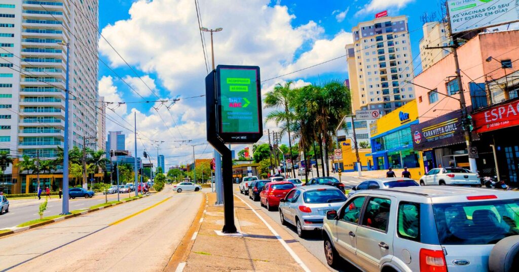 Imagem de uma rua com dois sentidos, no meio uma calçada com relógio/placa de painel de LED mostra publicidade visível mesmo sob sol intenso. 