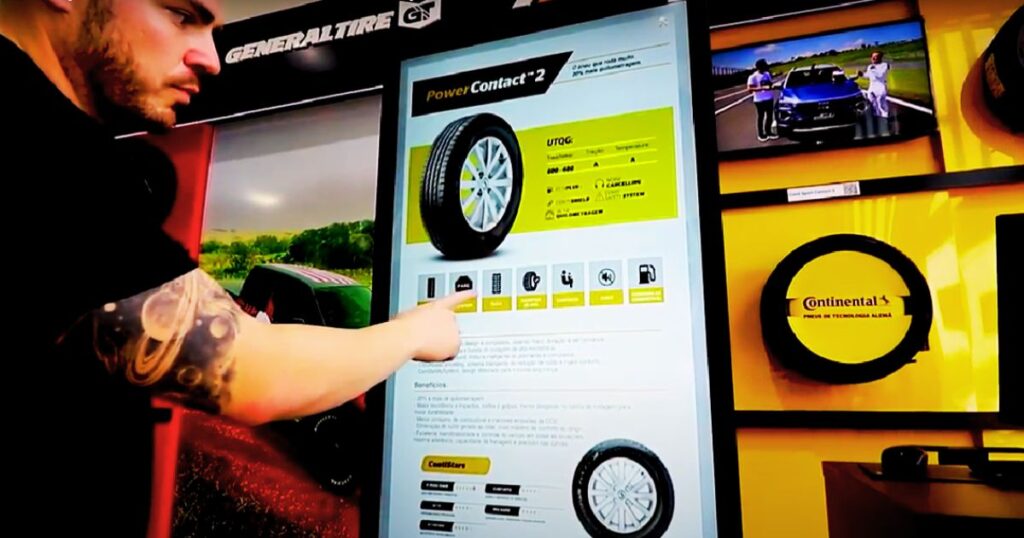 Imagem mostra um homem usando um sistema de tela touch de autoatendimento na escolha de pneus. Essa imagem serve para exemplificar como a tecnologia pode ser automatizada para melhorar a experiência do consumidor, sendo esta a maior das tendências do varejo em 2024.